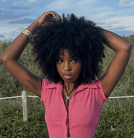 Woman with an afro in a pink sleeveless top standing outdoors with grass and sky in the background